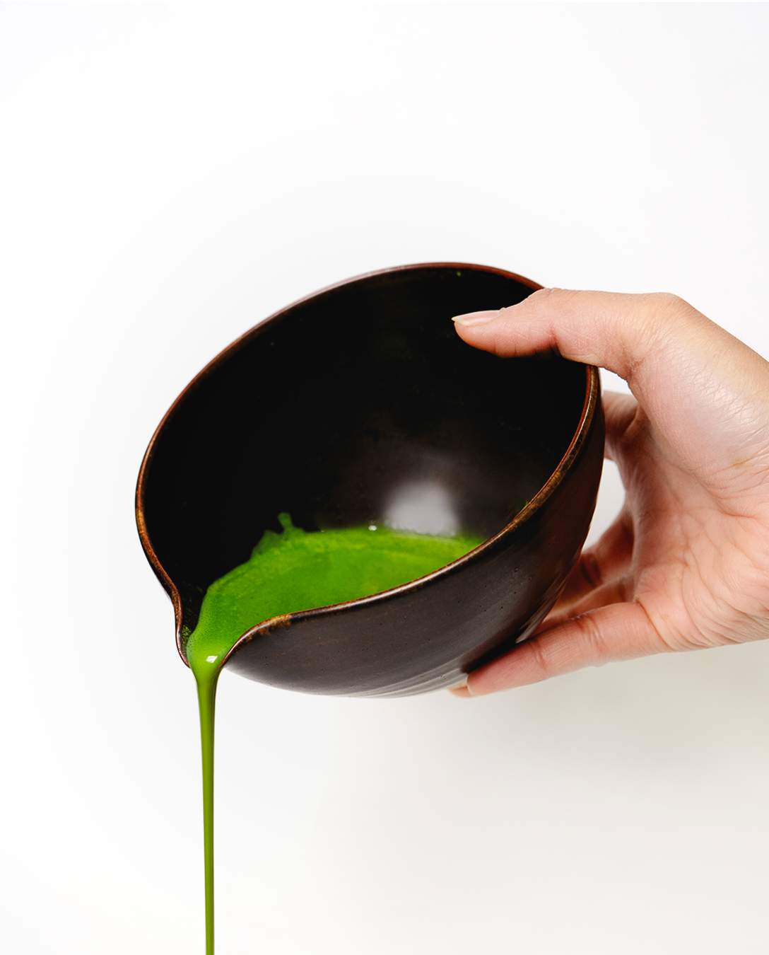 Hand holding a black bowl with Matcha pouring out on a white background